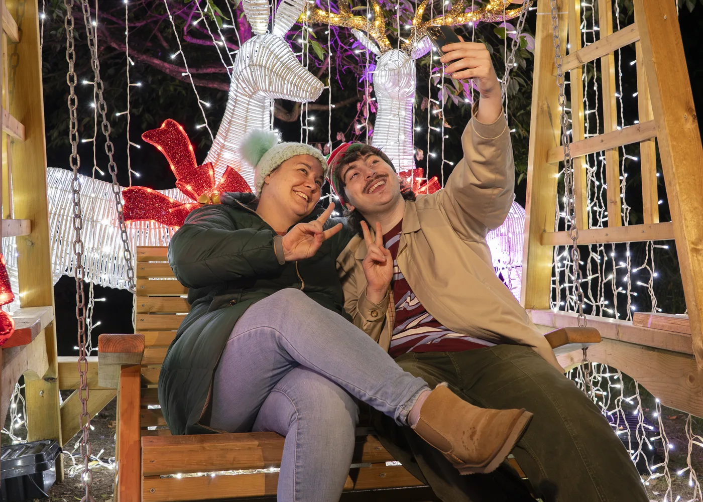 Two visitors take a selfie on a wooden swing seat hung with fairy lights, with light-up reindeer behind